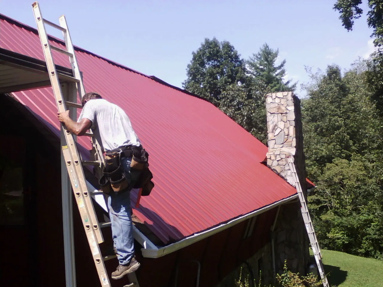 Roofer on a ladder beside a red metal roof and stone chimney — Soffit & Fascia Repair by Roofing Brothers in West Chester